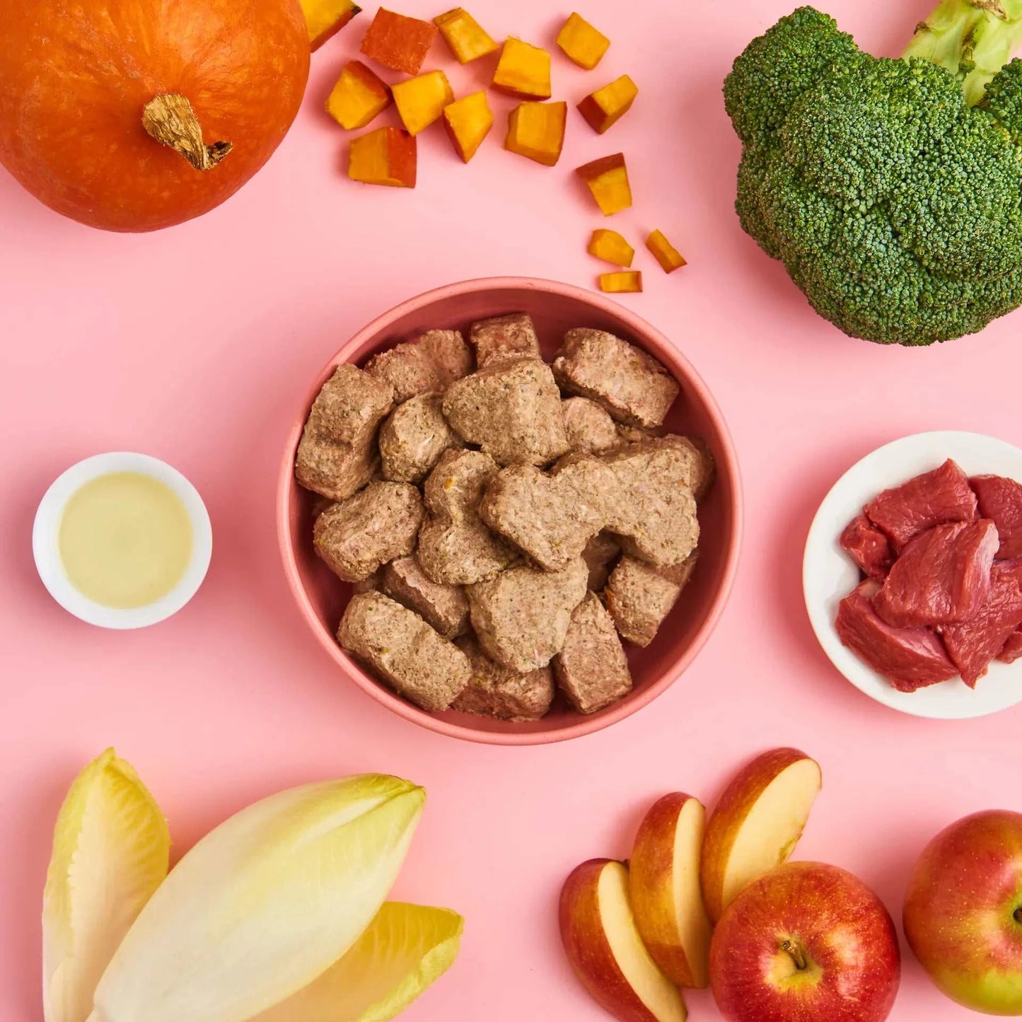 All-in-One Hundefutter mit Rindfleisch, Kürbis, Chicorée, Brokkoli und Apfel in einem Napf auf pinkem Hintergrund, umgeben von frischen Zutaten.