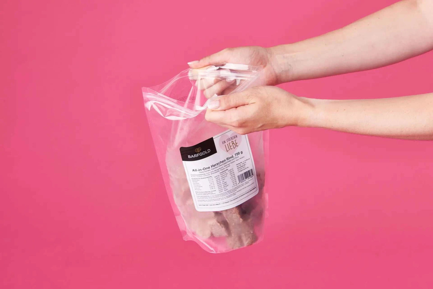 Person hält transparenten All-in-One Herzchen Rind 750 g Beutel vor pinkem Hintergrund.