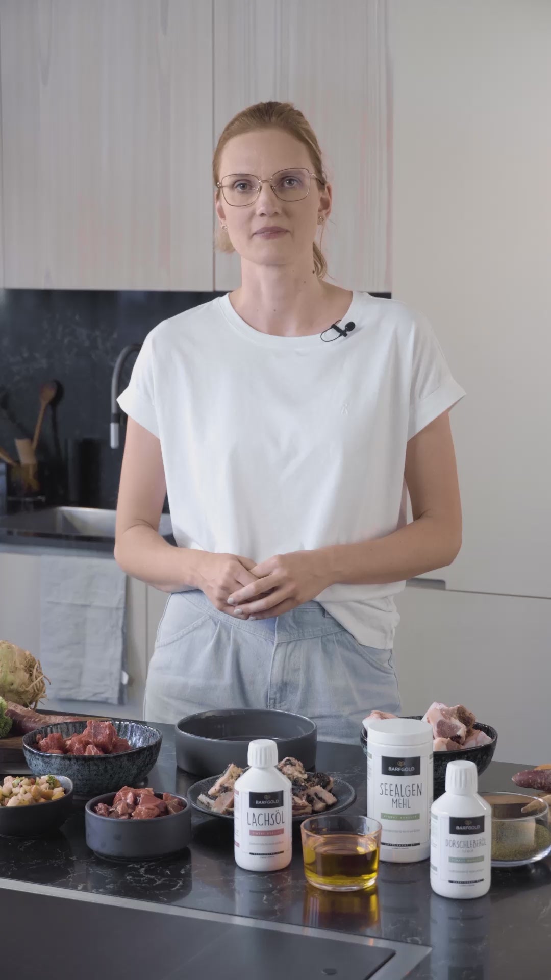 Frau mit weißem Top und Brille steht hinter einem Küchentresen. Auf diesem stehen sämtliche Produkte aus dem Shop. Sie erklärt wie BARF Ernährung funktioniert.
